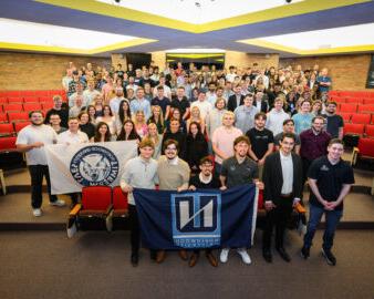 A large group of students gathers in a lecture hall, proudly holding a 竞技宝app下载 University flag and a university seal flag, united by their mission to stay connected.