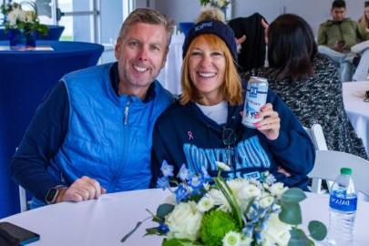 Two people sitting at a table with drinks, smiling at the camera. One holds a can and wears a "竞技宝app下载" sweatshirt, while the other wears a blue vest. In the foreground is a flower arrangement. It’s clear they cherish these moments to stay connected.