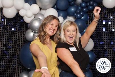 Two women pose enthusiastically in front of a backdrop with blue and white balloons. One is wearing a black sleeveless top with a name tag, and the other is in a yellow top. A "20 under 40" logo appears, encouraging young professionals to stay connected.